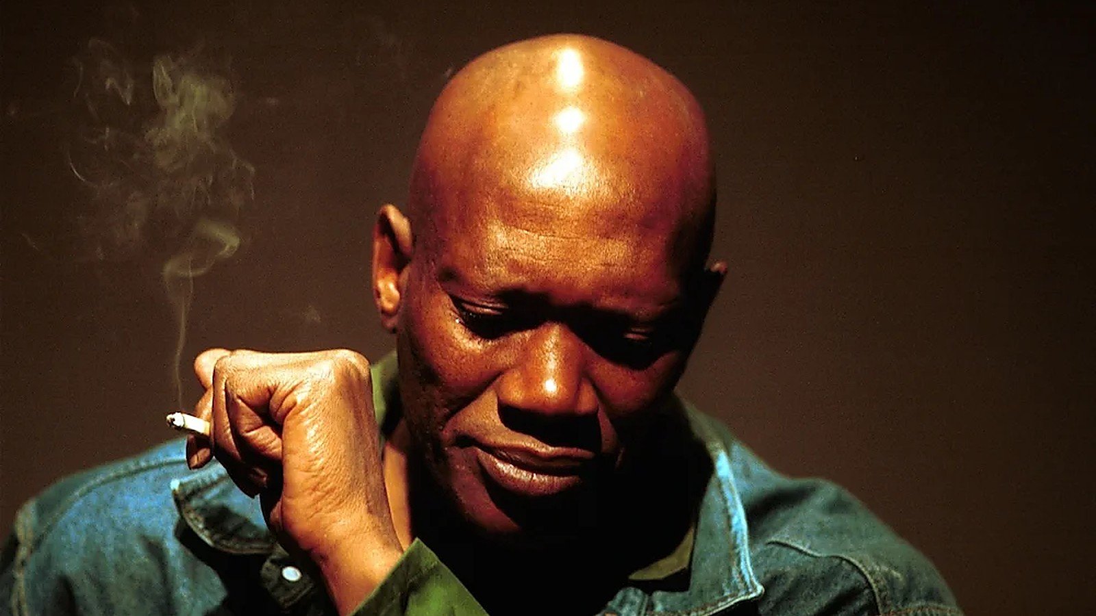 Duma Khumalo, head lowered and a cigarette in his hand, photographed against a dark ground.